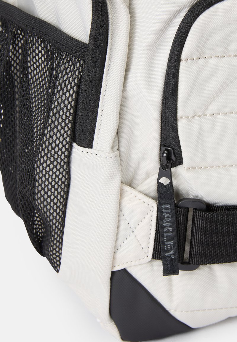 Backpack featuring a cream fabric body, black mesh pocket, black strap with hardware, and side label with logo detail.