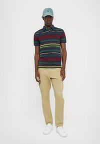Cămașă polo cu dungi roșii, verzi și galbene, asociată cu pantaloni cargo bej. Adidași albi și o șapcă albastră deschisă completează ținuta.
