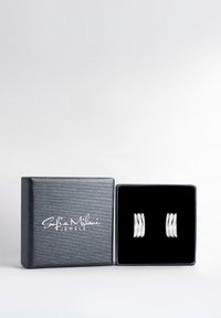 Silver ringformade örhängen visade i en svart låda fodrad med sammet och med en svart texturerad utsida. Lådan har varumärkesnamnet i vitt.