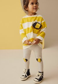 Gelb-weiß gestreiftes Langarmhemd mit Bienen-Pocket-Design, kombiniert mit grauen Leggings mit Bienen-Patches an den Knien und schwarzen Sneakers.