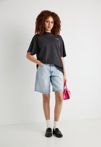 Γυναίκα με σγουρά μαλλιά που φοράει μαύρο υπερμεγέθες μπλουζάκι, ανοιχτόχρωμα τζιν σορτς, μαύρα μοκασίνια με λευκές κάλτσες, κρατώντας μια μικρή ροζ τσάντα.