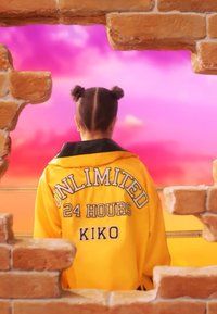 Persona con un peinado de doble moño, vistiendo una chaqueta amarilla con "UNLIMITED 24 HOURS KIKO" en la parte posterior, enmarcada por un muro de ladrillos roto.