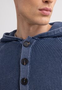 Mėlyna gobtuvė turinti tekstūruotą trikotažo raštą, tris juodus mygtukus ir šonkačių apykaklę. Artima žiūra, išryškinanti medžiagos detales.