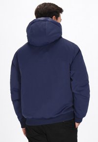 Sötétkék kapucnis dzseki sima textúrával, rugalmas mandzsettákkal és testhezálló aljjal. Egyszerű dizájn, látható minták vagy fémrészek nélkül.