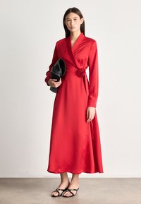 Czerwona satynowa sukienka zapinana z długimi rękawami, z różą w talii. Modelka trzyma czarną kopertówkę, nosząc proste czarne sandały.