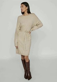 Pledukčio spalvos suknelė su ilgomis rankovėmis ir reguliuojamu liemeniu. Yra laisvo silueto ir glotnios tekstūros, derinama su ruda ilgaauliais batais.