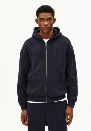 Junger Mann trägt einen marineblauen Kapuzenpullover mit Reißverschluss und Kordeln sowie passende Hosen vor einem schlichten weißen Hintergrund.