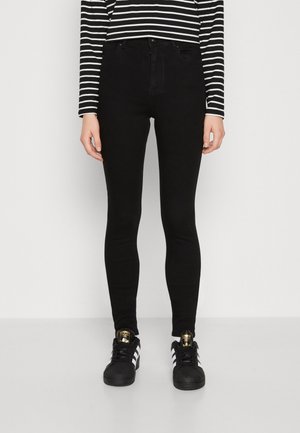 Person iført sorte skinny jeans, sort og hvid stribet langærmet top og sorte Adidas sneakers med hvide striber og guldlogoer.