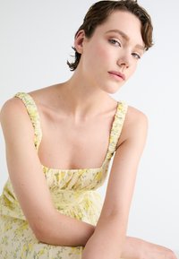 Žlté kvetinové šaty s nariasenými ramienkami, elastickými detailami a ľahkým materiálom. Majú štvorcový výstrih a splývavú sukňu.