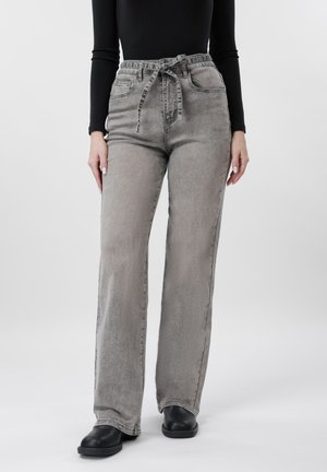 Žena u sivim širokim trapericama visokog struka, izblijedjelim, s povezanom trakom, crnoj dugoj majici s rukavima i crnim cipelama stoji ispred bijele pozadine.