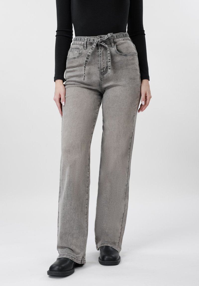 Nő magasderekú, kifakult szürke, bőszárú farmert visel, amelyhez kötött öv tartozik, fekete hosszú ujjú felsőben és fekete cipőben áll fehér háttér előtt.