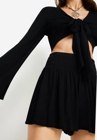 Μαύρο κοντό τοπ με μακριά μανίκια και λεπτομέρεια μπροστινού δεσίματος, συνδυασμένο με μαύρη φούστα με λάστιχο και φαρδιά σιλουέτα.