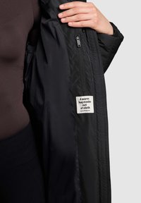 Svart isolerad jacka med ett slätt tyg, utrustad med en sidodragkedja och en etikett inuti med texten "En varm kram gjord av tyg".