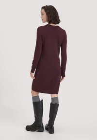 Bordeauxfarbenes Strickkleid mit langen Ärmeln, geripptem Halsausschnitt und figurbetontem Schnitt, kombiniert mit knielangen schwarzen Lederstiefeln und grauen Socken.