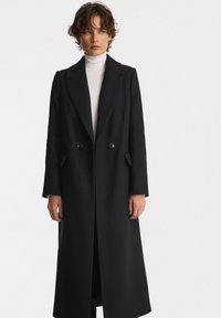 Czarny płaszcz dwurzędowy z dużymi klapami, bocznymi kieszeniami i prostym krojem, wykonany z teksturowanej tkaniny, sięgający poniżej kolan.
