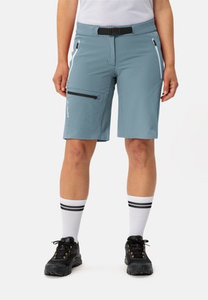 Himmelblaue, knielange Shorts aus elastischem Material, ausgestattet mit zwei Seitentaschen und einer Reißverschlusstasche, kombiniert mit weißen Socken und schwarzen Schuhen.
