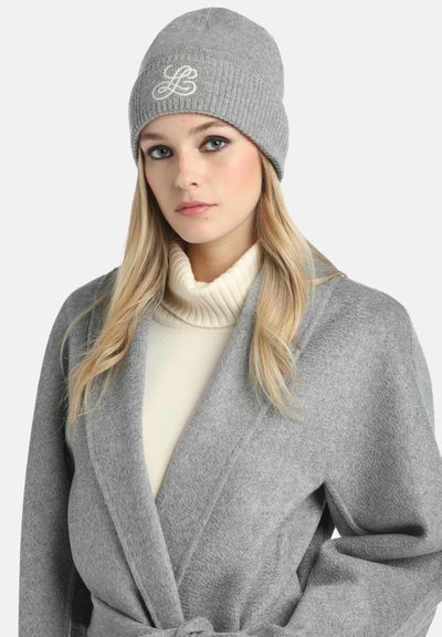 Jauna moteris, vilkinti pilką mezginį kepurėlę su logotipu, pilką paltą su diržu ir sviestinę turtleneck megztinį, žiūri šiek tiek į šoną.