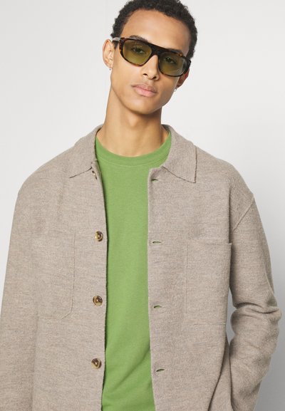 Jachetă din lână bej, cu guler și închidere cu nasturi, purtată peste un tricou verde. Modelul poartă ochelari de soare din tortoise dark cu lentile verzi.