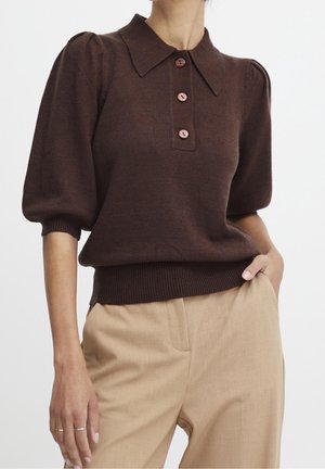 Brązowy dzianinowy sweter polo z kołnierzykiem, krótkimi bufkami, czterema różowymi guzikami i ściągaczem u dołu. Połączony z beżowymi spodniami.