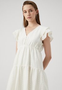 White dress featuring a V-neck, short ruffled sleeves, and a tiered skirt with textured patterns and button accents down the front.