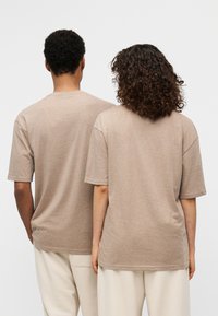 Μπεζ T-shirts με κοντά μανίκια σε χαλαρή εφαρμογή, κατασκευασμένα από μαλακό βαμβακερό μείγμα. Ακατέργαστες άκρες στο τελείωμα και τα μανίκια. Φορεμένα με ανοιχτόχρωμο παντελόνι.