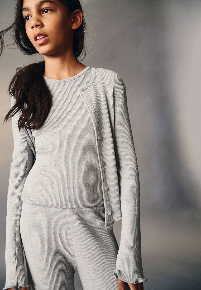 Jeune personne vêtue d'un haut à manches longues en maille côtelée gris clair, d'un cardigan assorti avec des boutons et d'un pantalon, sur un fond neutre uni.