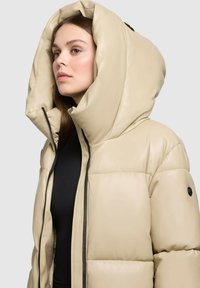 Chaqueta acolchada beige con una gran capucha, presenta un cierre de cremallera en la parte delantera, paneles cosidos y detalles en negro. Textura suave y brillante.