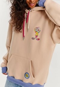Béžová mikina s ružovými a bielymi šnúrkami, prednou vreckom a grafikou karikatúry. Mäkka tkanina, zakrivený lem a fialové manžety.