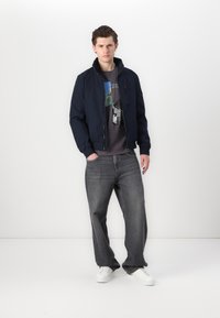 Giubbotto bomber navy con colletto alto, t-shirt grigia con grafica, jeans grigi larghi e sneakers bianche su uno sfondo semplice.
