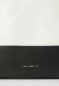 Crna kožna torba s bijelim gornjim dijelom, koja ima glatku teksturu i srebrni logo "KARL LAGERFELD" na prednjoj strani. Pravokutnog je oblika.