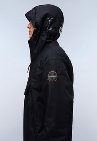 Person, die eine schwarze Kapuzenjacke mit einem runden Logo-Patch am Oberarm trägt, blickt nach links vor einem einfarbigen hellen Hintergrund.