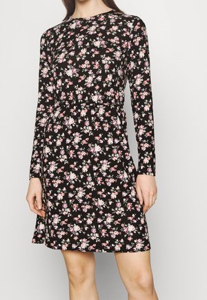 Kvinde iført en sort langærmet kjole med et pink og hvidt blomstermønster, tætsiddende ved taljen og afsluttende over knæene.
