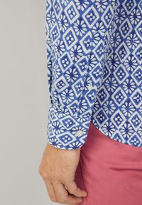 Camisa de patrón azul y blanco con un diseño geométrico y acentos florales, hecha de tela, con puños abotonados y un dobladillo curvado.