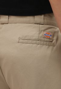 Bežne bombažne twill hlače z zadnjim žepom, ki vsebujejo šivano oznako zlogom "Dickies" v rdeči in modri barvi.