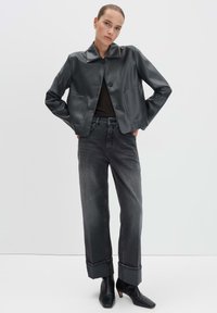 Schwarze Leder-Cropped-Jacke mit reverskragen, zwei vorderen Taschen und Knopfverschluss, kombiniert mit hochgeschnittenen grauen Jeans und schwarzen Absatzstiefeletten.