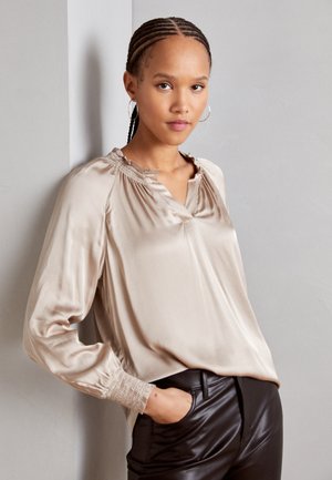 Satin beige bluse med V-hals, samlede halsudskæring og lange ærmer med smockede manchetter, parret med sorte læderbukser.