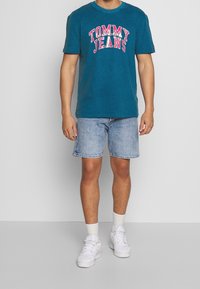 Camiseta de algodón en color azul verdoso con "TOMMY JEANS" en letras rosas, combinada con shorts de mezclilla claros y zapatillas blancas, de pie frente a un fondo neutro.