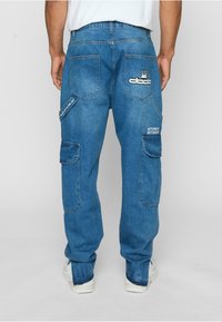 Blaue Denim-Cargo-Hosen mit mehreren Taschen, gestickten Logos und einer lockeren Passform. Sie verfügen über einen strukturierten Stoff und sichtbare Nähte.
