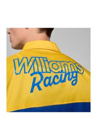 Żółta kurtka z teksturowanej tkaniny z niebieskim napisem "Williams Racing" na plecach. Zawiera kołnierz oraz poziome niebieskie paski.