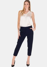 Giovane donna in piedi con una mano nella tasca dei pantaloni navy, indossando una blusa bianca senza maniche e tacchi a punta neri, su sfondo semplice.