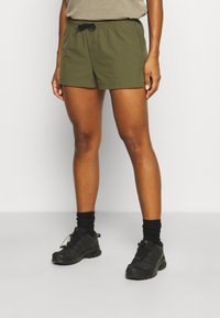 Shorts verts avec une taille élastique et un cordon de serrage. Comprend des poches latérales et un logo sur la cuisse gauche. Portés avec des chaussures de randonnée noires.