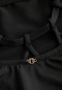 Schwarzer Stoff mit Netzakzenten und glatter Textur. Elastische Träger mit Zickzacknaht und einem goldenen runden Verschlussdetail am Boden.