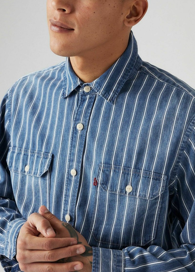 Homme portant une chemise en denim à rayures verticales bleues et blanches avec deux poches poitrine, mains jointes montrant un tatouage sur la main droite.