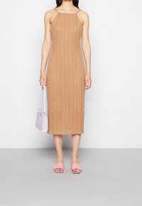 Nainen, joka on pukeutunut hihattomaan vaaleanbeigeen neulemidimekkoon, pitelee pientä laventelinväristä käsilaukkua ja hänellä on jalassaan vaaleanpunaiset avokkaat, seisoo harmaalla lattialla.