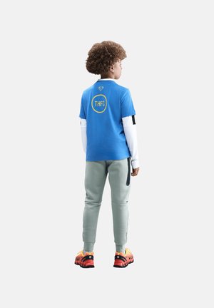 Barn med krøllet hår står med ryggen til iført en blå THFC-trøje over en hvid langærmet trøje, grå bukser og orange og sorte sneakers.