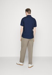 Uomo di spalle che indossa una camicia corta blu navy, pantaloni marrone chiaro e sneakers bianche, su uno sfondo chiaro e semplice.