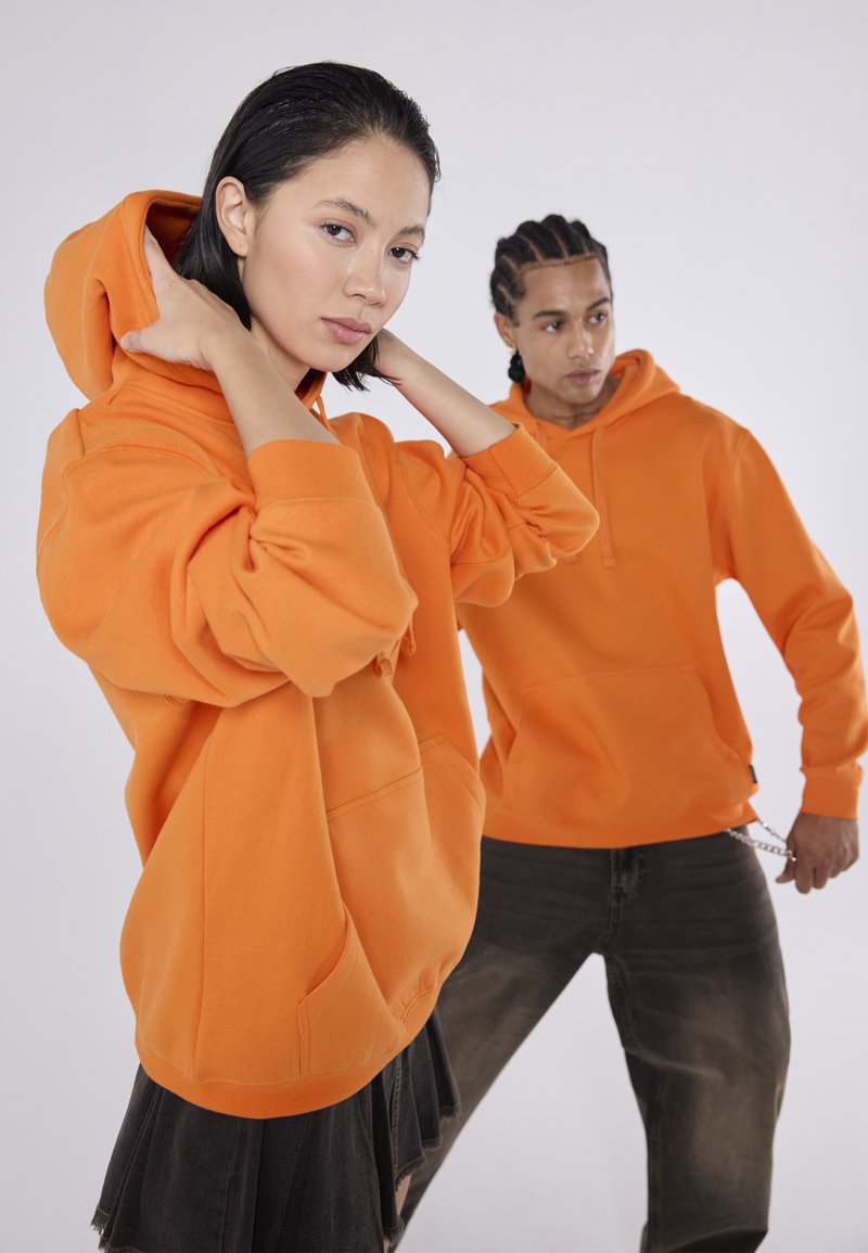 Deux individus portent de grands sweats à capuche orange en tissu doux. La femme a les cheveux courts et l'homme arbore des cheveux tressés.