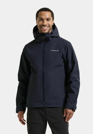 Smiling man wears a black zippered Didriksons jacket with a hood, standing against a plain light background.