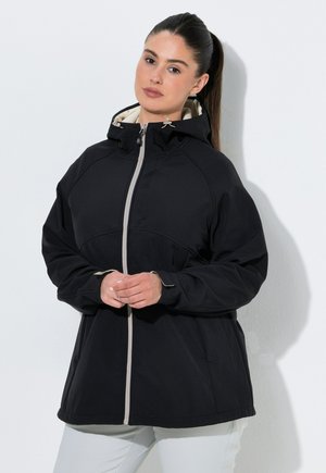 Frau mit dunklen Haaren im Pferdeschwanz, die eine schwarze Kapuzenjacke mit einem beigen Reißverschluss und weiße Hosen trägt, steht vor einer weißen Wand.