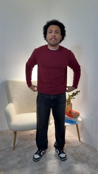 Μακρυμάνικο πουκάμισο σε μαρονοκόκκινο χρώμα, μαύρο τζιν και μαύρα αθλητικά παπούτσια με λευκές λεπτομέρειες. Στέκεται δίπλα σε μια λευκή, υφασμάτινη πολυθρόνα και βιβλία.